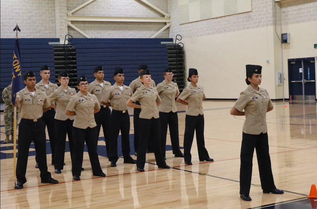 Stony Point High School NJROTC cadets standing in formation at parade rest on the gymnasium basketball court during a drill competition or inspection. The cadet commander stands in front leading the platoon, while a cadet on the left holds the Stony Point Navy JROTC flag.