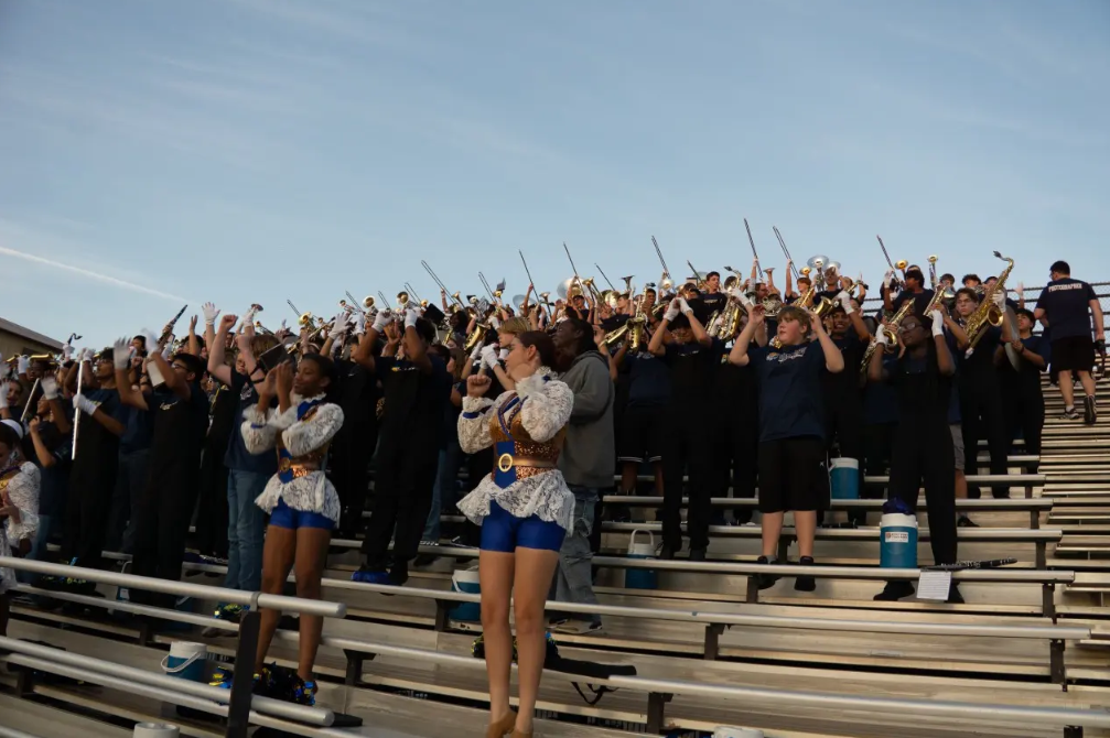 Wide-angle photograph of the Stony Point High School Tiger Marching Band and Color Guard filling the metal bleachers at Tiger Stadium. Color guard members in blue and white lace outfits stand in front while band members hold up brass and woodwind instruments performing stand tunes under a clear sky.