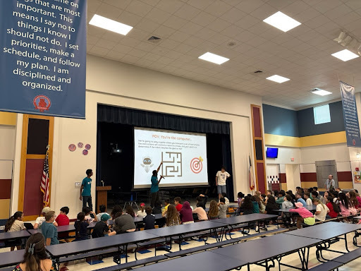 A wide cafeteria view showing students watching a presentation slide explaining step-by-step programming concepts.