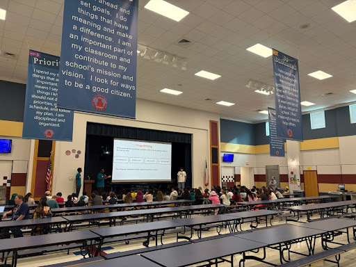 Students sit at cafeteria tables watching a UT Computer Science Roadshow slide about a robot maze game in the cafeteria.