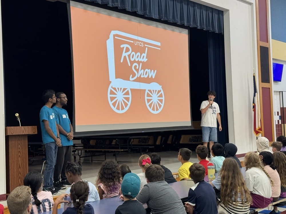 Three presenters stand on stage in front of a UTCS Road Show slide while elementary students listen attentively.