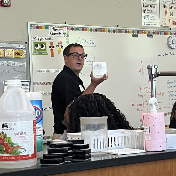 A man stands in front of a white board shows a tile with black finger prints to a class.