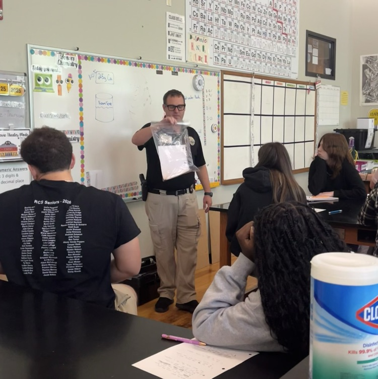 A man holding up a plastic bag with a piece of paper inside of it in front of a class