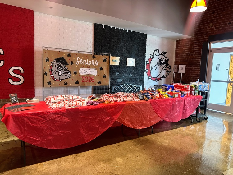 a table full of Chick-fil-A sandwiches, chips, and sodas with a red table cloths and a sign that say Class of 2026 behind it.
