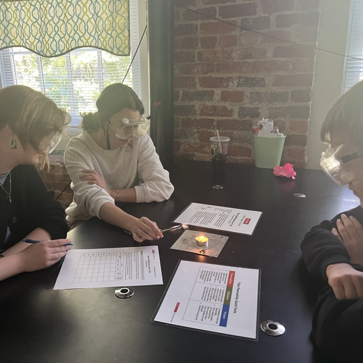 3 students sit at a lab table while one holds a fiber sample over a tea light candle with tweezers.
