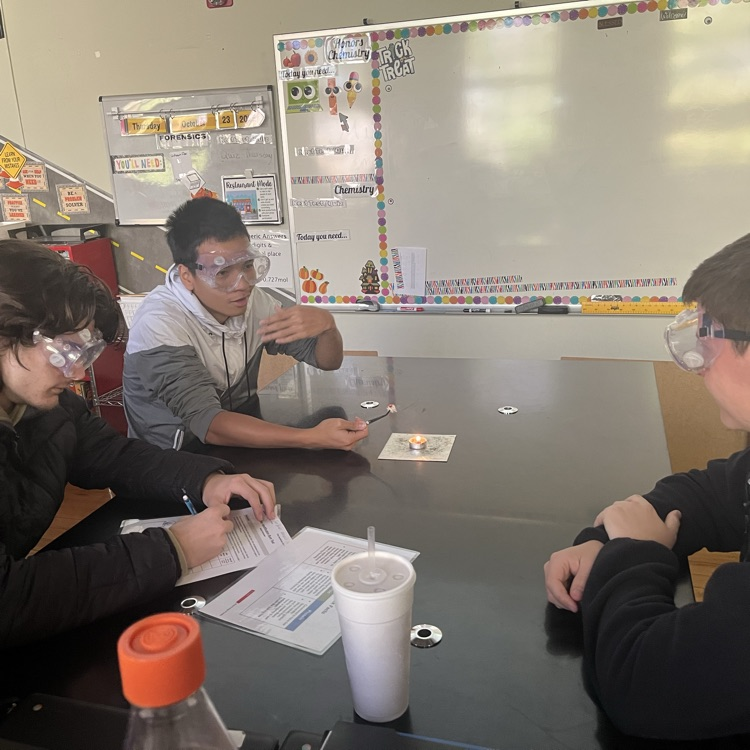 3 students sit at a lab table while one holds a fiber sample over a tea light candle with tweezers.