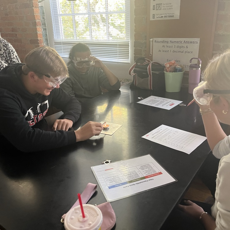 3 students sit at a lab table while one holds a fiber sample over a tea light candle with tweezers.