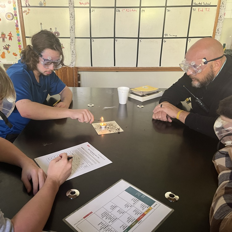 3 students and the principal sit at a lab table while one holds a fiber sample over a tea light candle with tweezers.