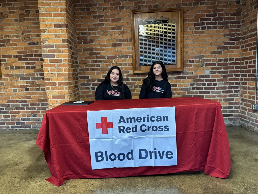 Two females sitting at a table with a red tablecloth and an American Red Cross Blood Drive sign on it. 