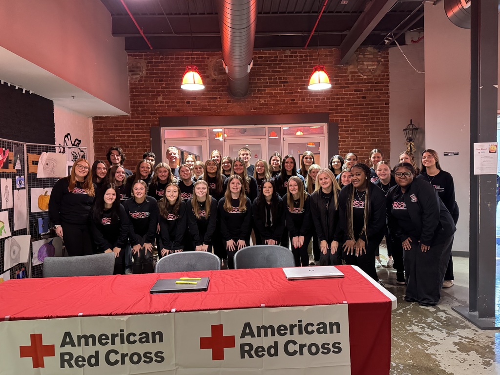 A large group of students posing in front of a table with a red table cloth and an American Red Cross sign on it. 