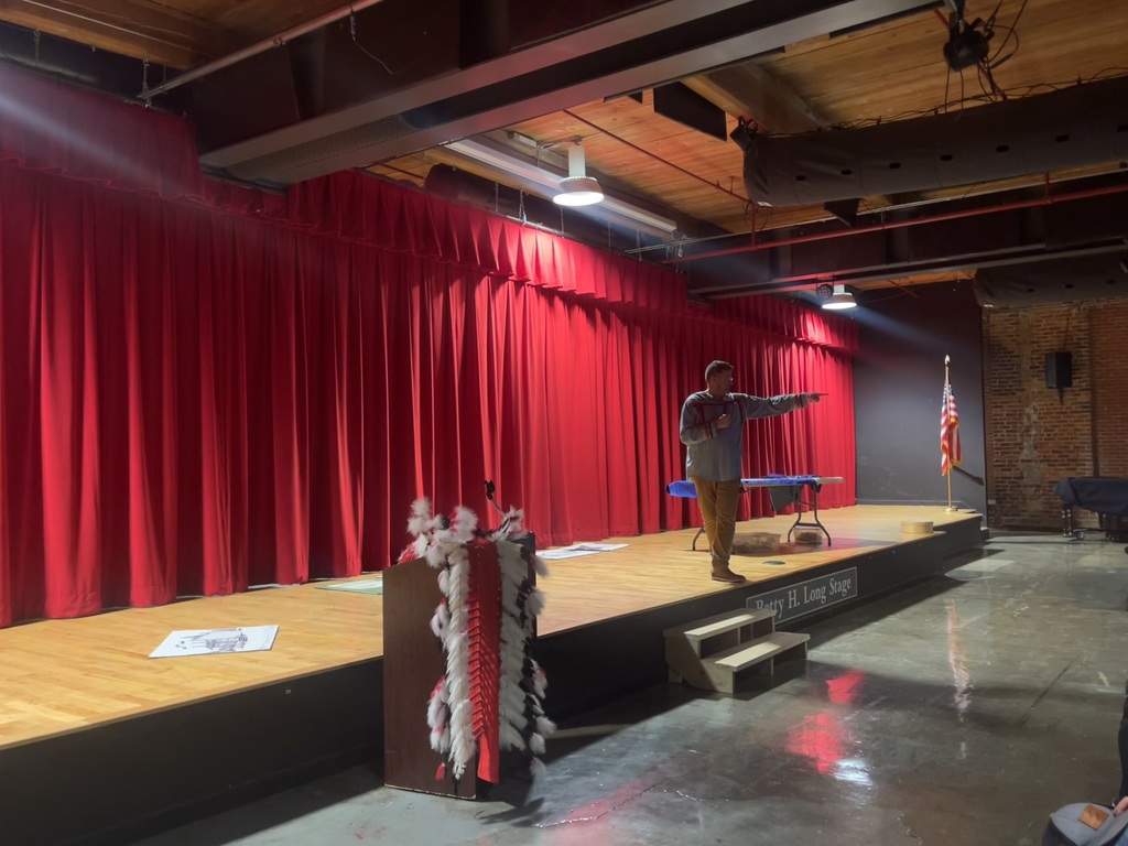 Will Paul on the MPR stage talking with a mic in hand and a traditional Indian headdress on a podium close to him. 