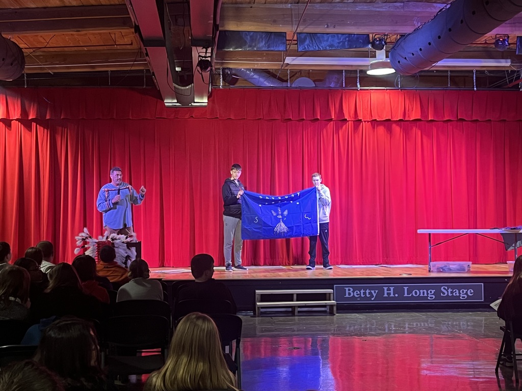 Will Paul on the MPR stage talking while two students hold a Sappony Tribe flag. 