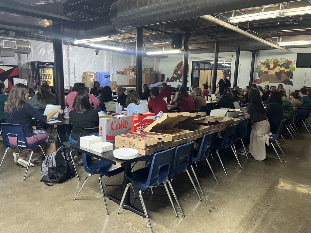 Boxes of pizza and soda with plates and napkins on a table in the dining hall with all the staff sitting at tables during a faculty meeting. 