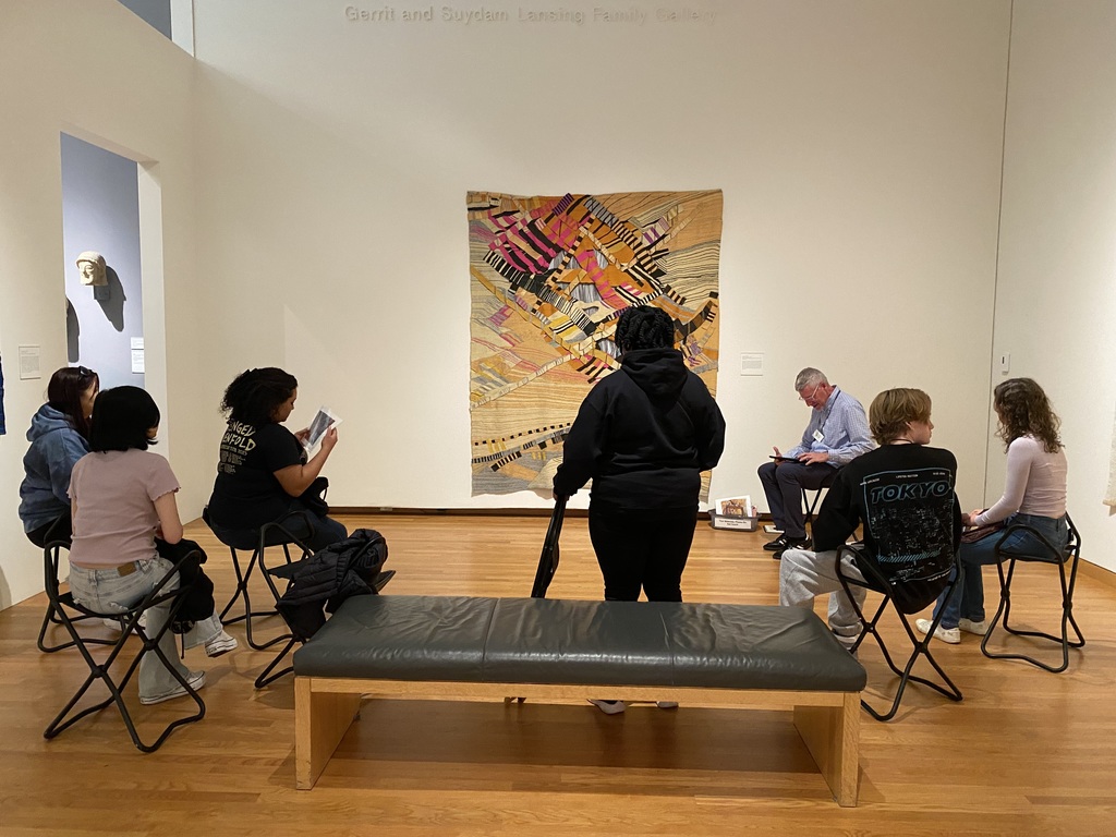 people sitting on foldable chairs in an art museum, in front of a large piece of art.