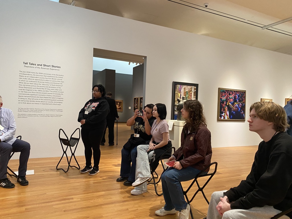 5 students sitting in foldable chairs, looking at art.