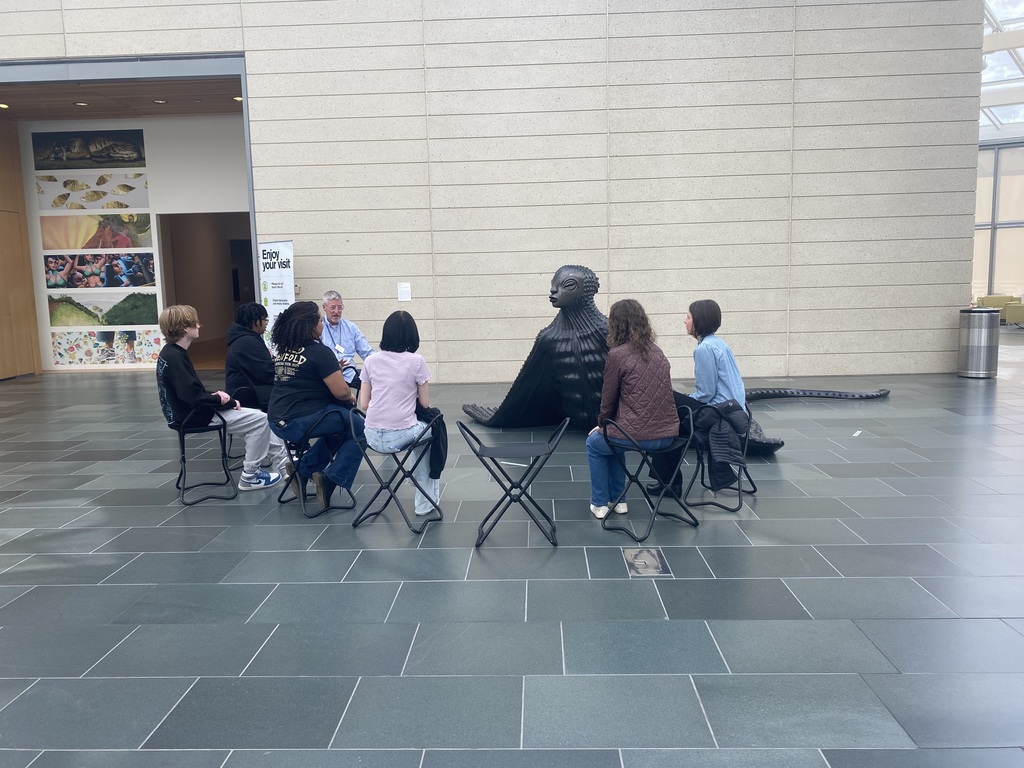 A group of people sit in foldable chairs listening to a man speak about art in front of a sculpture.