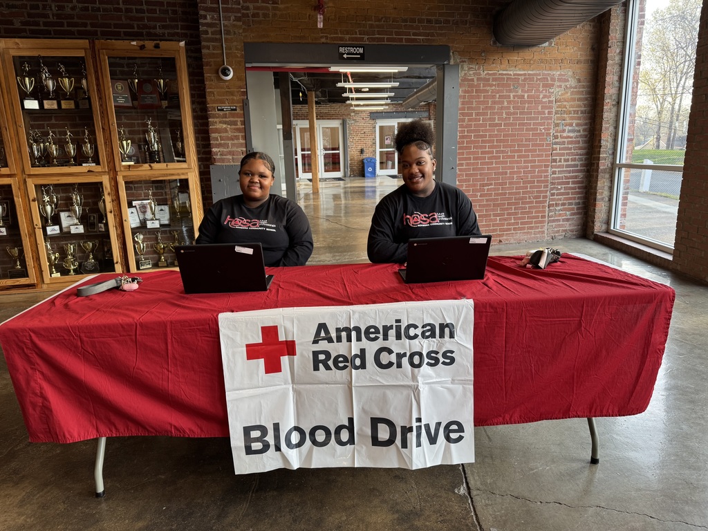 Two RCS students sitting at a table with a red table cloth with an American Red Cross Blood Drive sign on it.