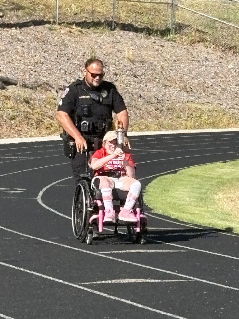 A young girl in a wheelchair carrying a torch with a police officer pushing her on a track outside