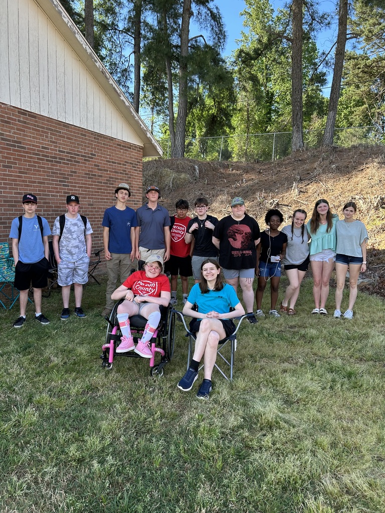 A group of students standing together, smiling outside with a girl in a wheelchair and another girl sitting in a chair in front of them. 