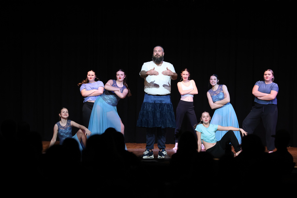 A group of teenage girls dance costumes posing around a male teacher wearing a white tshirt and a tutu