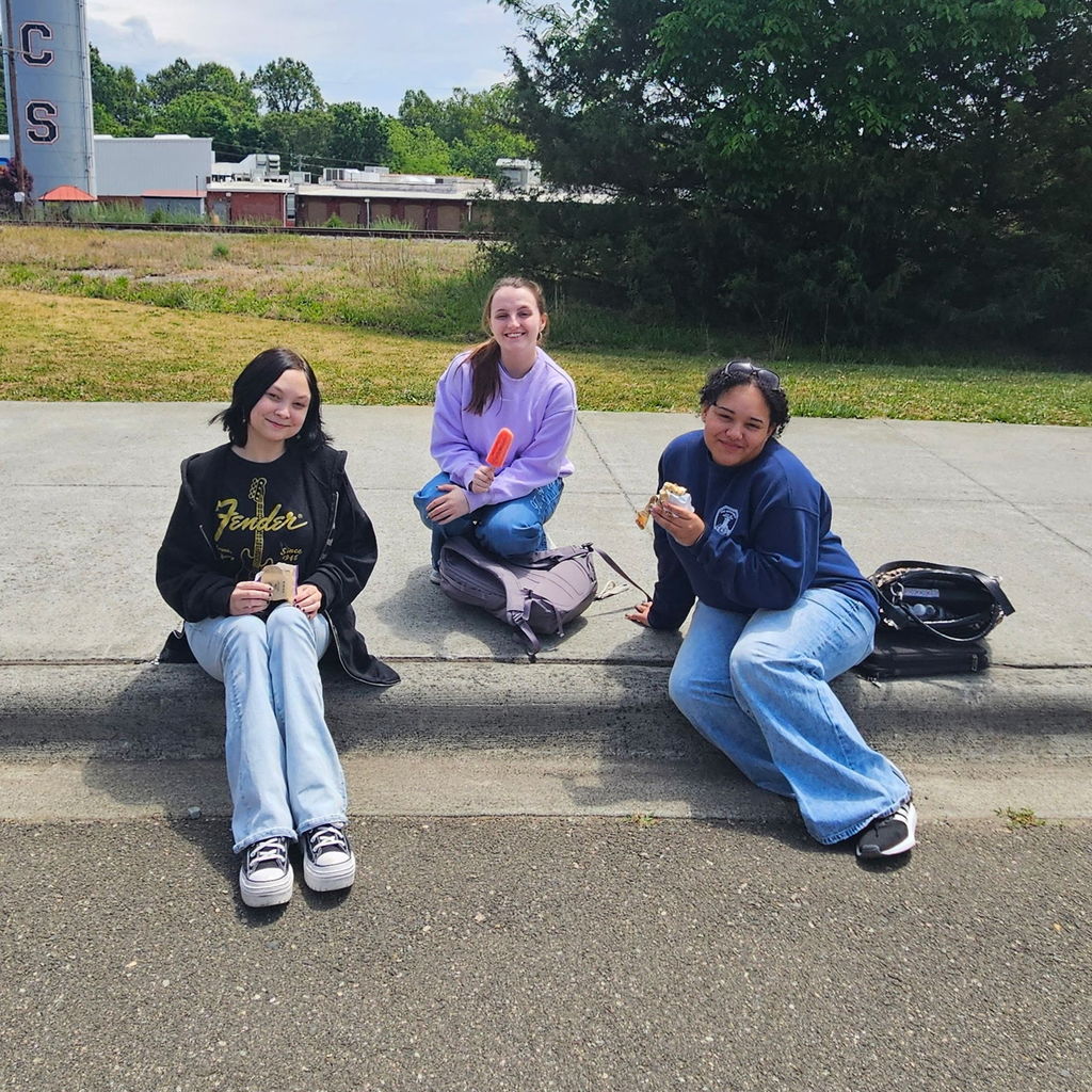 3 high school students smiling, sitting on a curb, holding ice cream or a popsicle. 