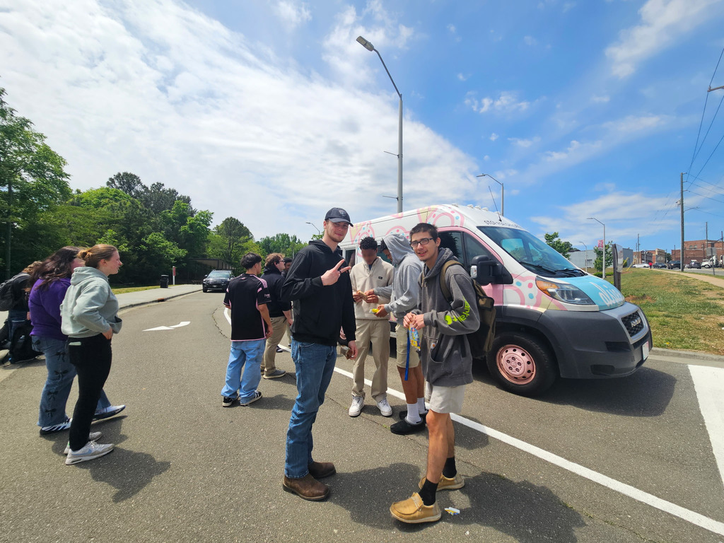 High school students standing in line for the ice cream van in a parking lot. Two are facing the camera smiling.