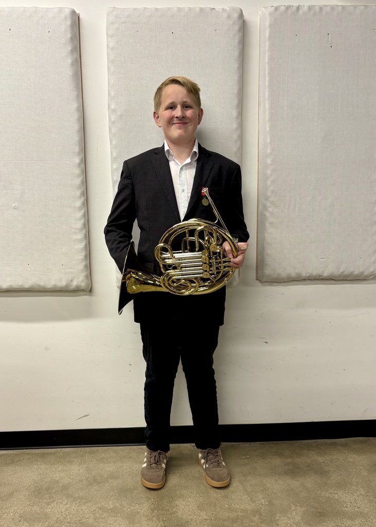 A teenage boy in a black suit coat and a white button down shirt, holding a french horn standing in front of a cream wall.