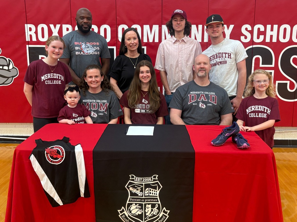 Kylie Barker and her family, friends, coach, and school administrator behind a table with a red table cloths and a black runner with a cross country jersey on one side and a pair of running shoes on the other. There are 10 people pictured