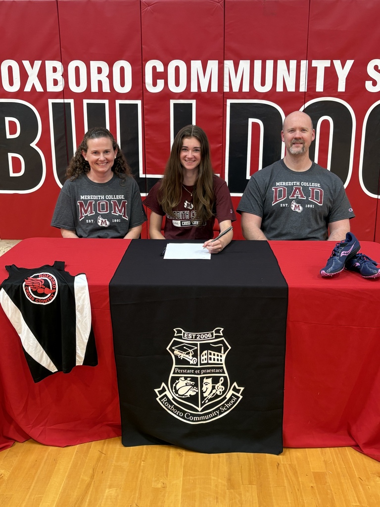 Kylie Barker and her parents sitting at a table with a red table cloth with a black runner in the middle of it. There is a cross country jersey on the left side of the table and a pair of running shoes on the right side. Kylie is in the middle holding a pen to a piece of paper.