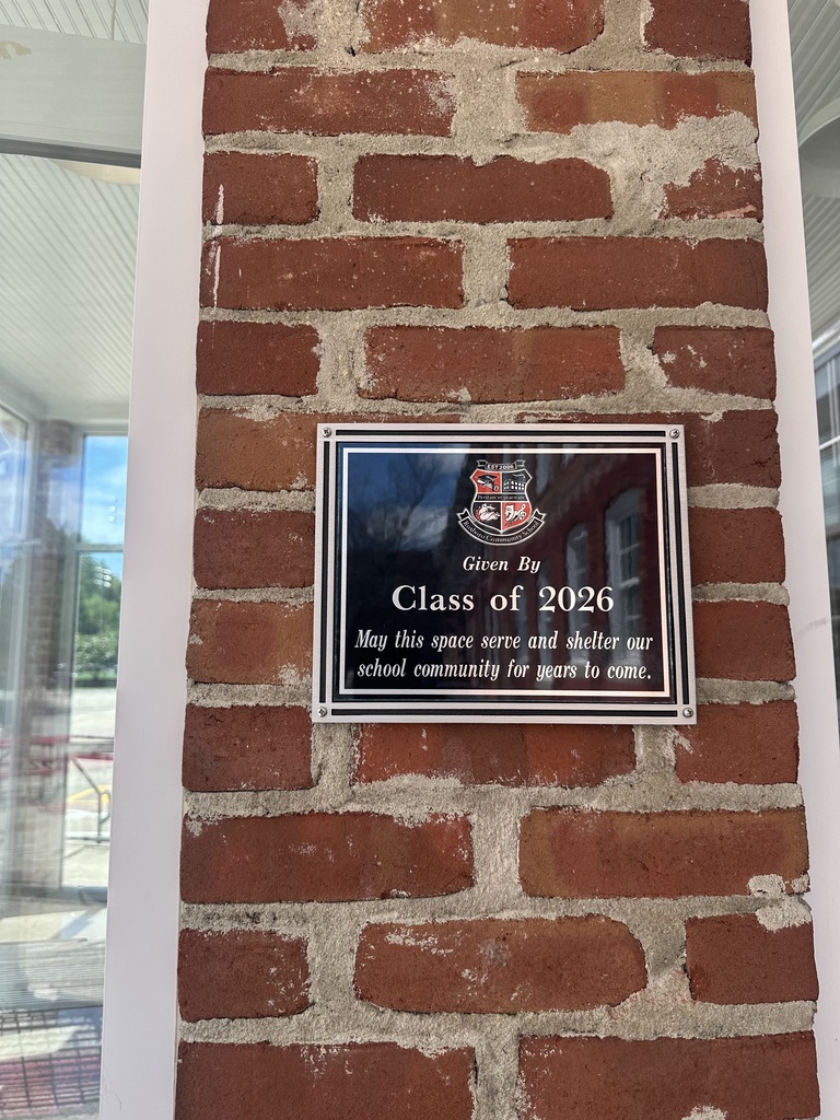 A black plaque with a silver border hung on a brick wall. The plaque has the RCS crest in the top middle with the following text underneath: Given By Class of 2026, May this space serve and shelter our school community for years to come. 