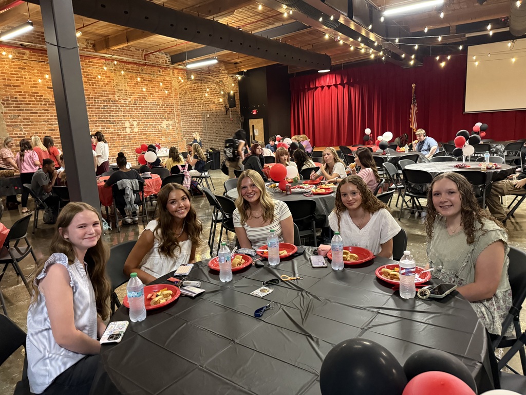 5 teenage girls smiling, sitting at a table with a black tablecloth and plates of food. 