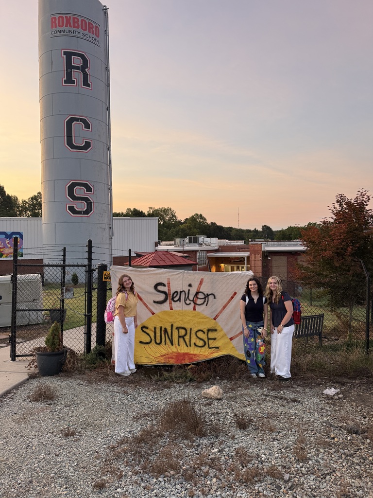 3 teenage girls standing outside of RCS, in front of a sign that says senior sunrise and with a sunshine behind it. 