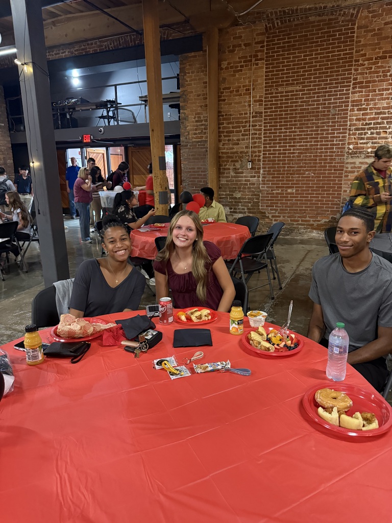 3 teenagers sitting at a table with a red tablecloth with plates of food
