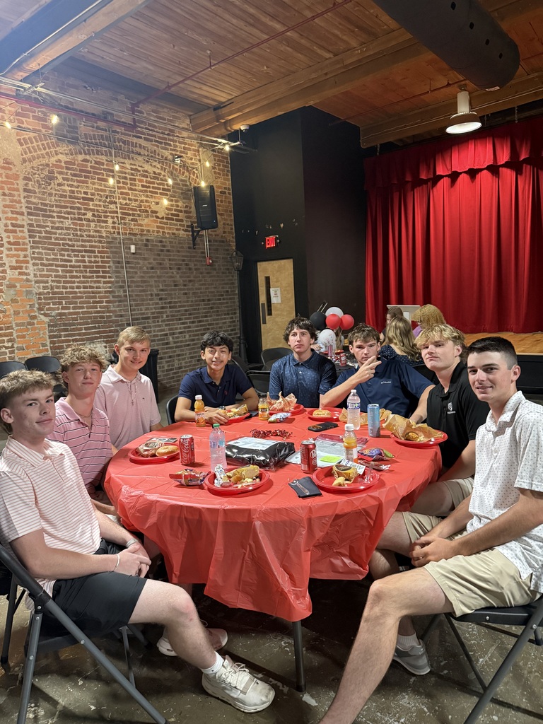 8 teenage boys sitting at a table with a red table cloth and plates of food 