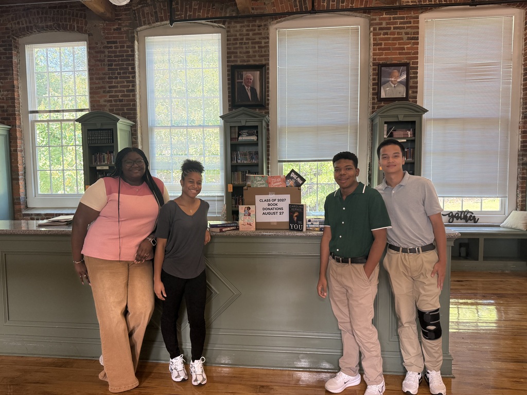 4 high school seniors (2 girls on the left, 2 boys on the right) standing in the RCS library with a box of books on the counter between them. 