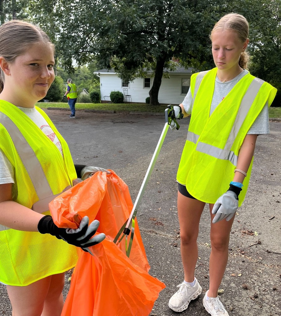 On girl. on the right puts trash in an orange trash bag using a grabber stick while another girl holds the bag open. Both are wearing yellow safety vests.