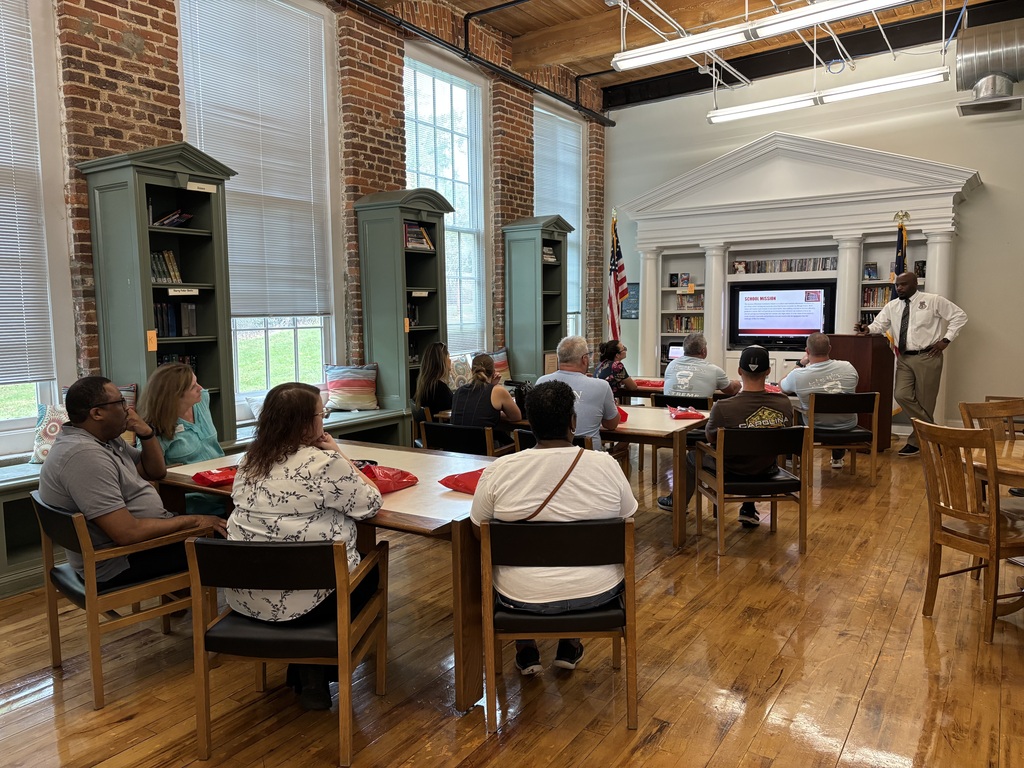 A group of adults sitting at tables, facing a TV with a slideshow on it and a man standing at a podium in the RCS library