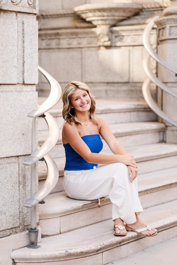 A photo of a girl wearing a blue tube top and white pants and white sandals sitting on beige steps. 