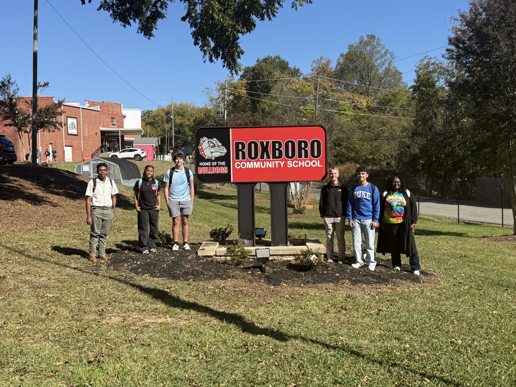 6 RCS seniors smiling with the red and black Roxboro Community Sign in the middle of them with new azaleas and mulch around it. 