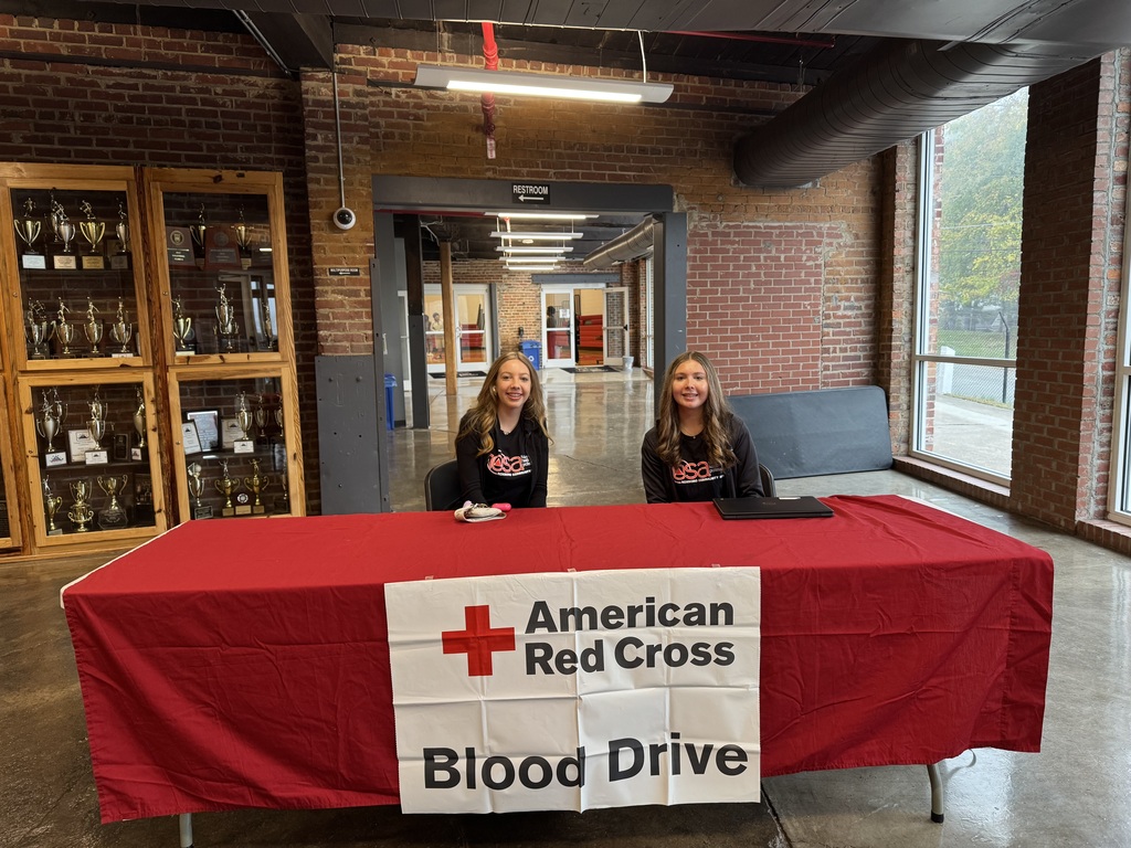 Two teenage girls sitting at a table with a red table cloth with a white sign that says "American Red Cross Blood Drive"