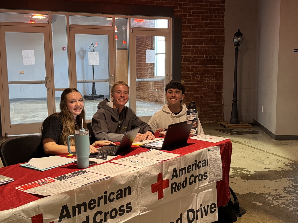 Three teenagers sitting at table for an American Red Cross Blood Drive with lapots in front of them. 