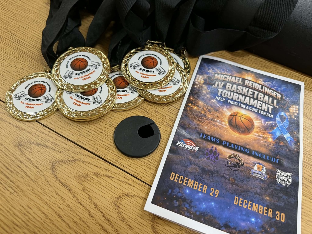 The image shows a wooden table with several gold basketball tournament medals stacked together on the right side. Each medal is attached to a black ribbon and features a basketball graphic with text referencing the Michael Reidlinger Memorial Tournament and Roxbury. On the left side of the table is a printed event program or flyer titled “Michael Reidlinger Memorial JV Basketball Tournament,” with dates listed as December 29 and December 30. The design includes a basketball image, school logos, and a blue ribbon symbol representing ALS awareness.