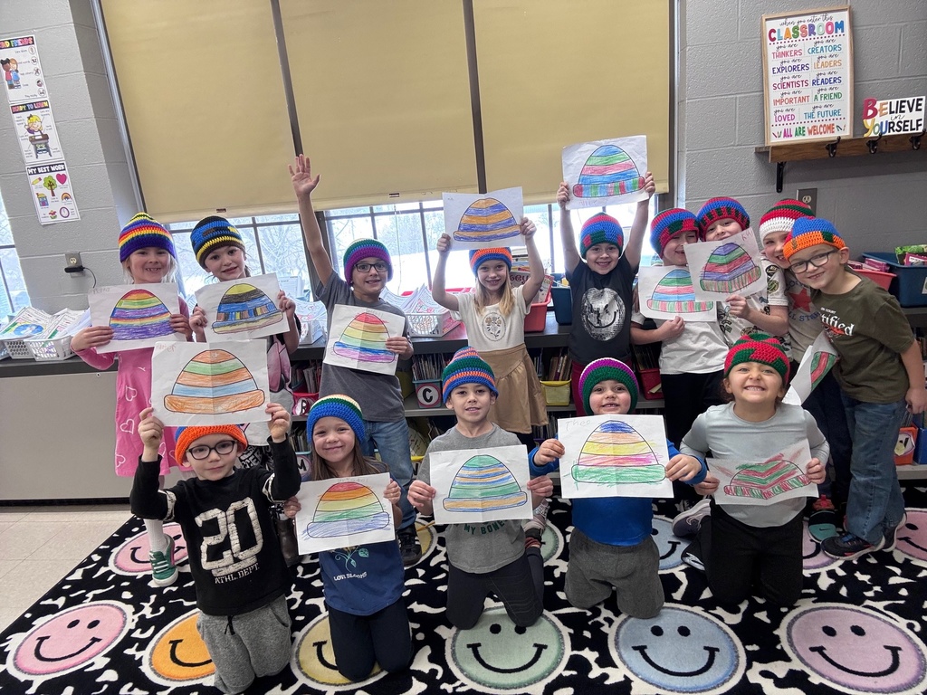 The photo shows a group of first-grade students standing and kneeling together inside a classroom. Each child is wearing a colorful crocheted hat with stripes in bright colors such as blue, green, red, yellow, and purple. The students are holding up drawings of hats that match the crocheted ones they are wearing. The classroom background includes shelves, posters, and windows, and the students are standing on a rug decorated with large smiley faces. Many of the children are smiling and raising their artwork proudly, showing the connection between their original designs and the finished hats.