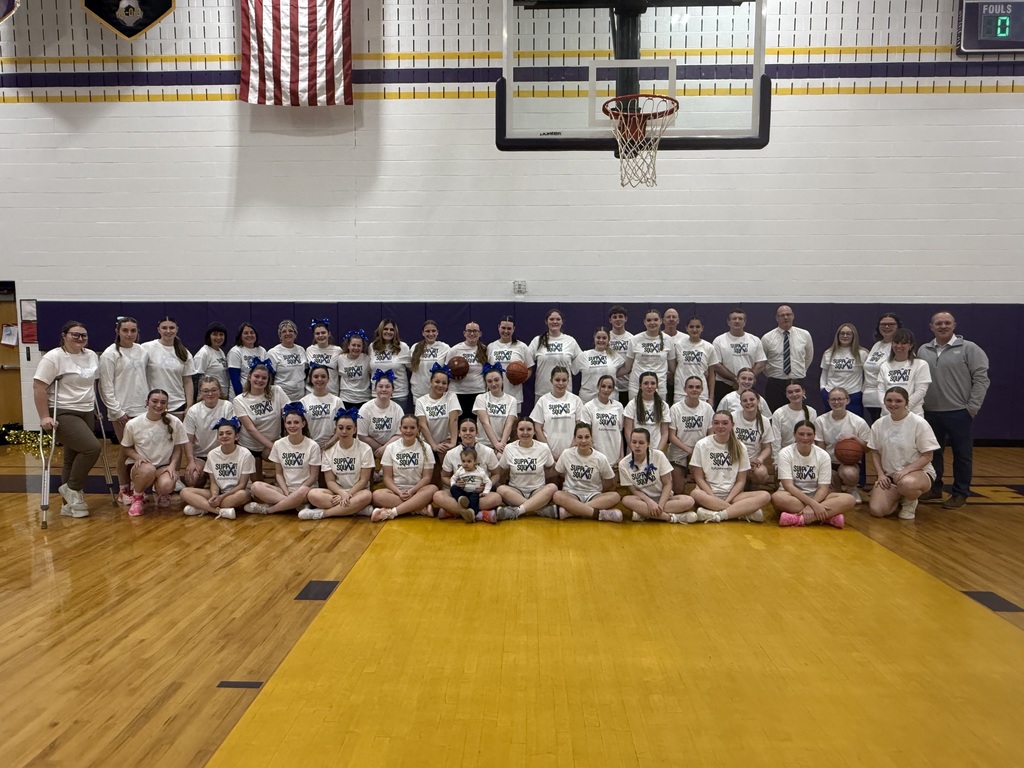 The image shows a large group photo taken inside a school gymnasium. All of the basketball teams that participated in the event are seated and standing together on the gym floor, along with their coaches and cheerleaders from GCR, Cherry Valley, and Schenevus. The group is arranged in several rows, with students sitting cross-legged in the front and others standing behind them. Most of the students are wearing matching white T-shirts that say “Support Squad,” while many of the cheerleaders have blue bows in their hair, matching the Blue Out theme.  A basketball hoop and backboard are visible above the group, and an American flag hangs on the wall in the background. The gym floor is wooden with a large yellow section in the foreground. Coaches and staff stand at the sides and in the back, dressed in professional or school attire. The overall mood of the photo is positive and celebratory, showing a united group of athletes, cheerleaders, and coaches coming together to support ALS research.
