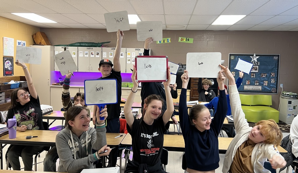 The image shows a classroom full of middle school students seated at desks. Many students are smiling and raising small whiteboards or dry-erase boards above their heads. Most of the boards have the word “left” written on them. The students appear excited and engaged, with several laughing and leaning toward each other. The classroom has cabinets along the back wall, colorful decorations, and posters. The overall mood is energetic and joyful, showing students proudly sharing their answers during a class activity.