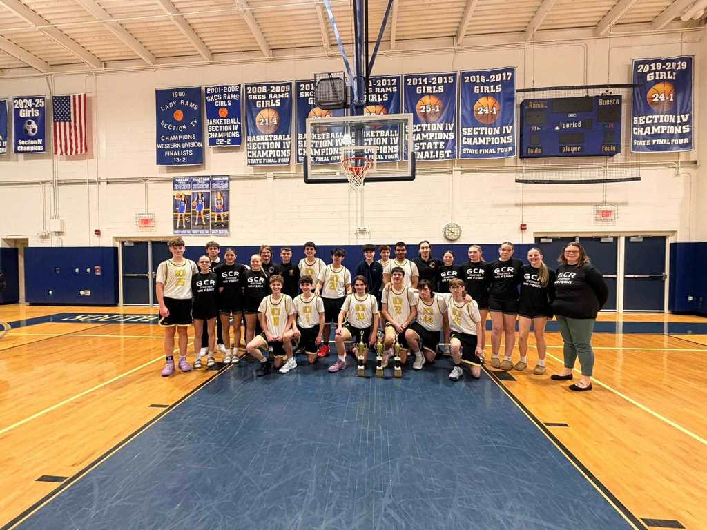 This image shows a large group of high school basketball players, coaches, and team members gathered on a gymnasium court for a celebratory photo. The group is centered beneath a basketball hoop and backboard.  In the front row, several boys wearing white GCR jerseys with gold numbers kneel on the blue-painted center section of the court. Three gold championship trophies are placed on the floor directly in front of them. The players are smiling and leaning slightly toward one another.  Behind them stands a larger group of teammates and supporters. Some wear white game jerseys, while others wear black shirts with “GCR” printed in gold lettering. A few adult coaches stand at the ends of the group. Everyone appears relaxed and proud, posing after a successful season.  The gym has a polished wooden floor with blue boundary lines. Blue championship banners hang high on the cream-colored walls in the background, listing past Section IV titles and seasons. An American flag hangs to the left, and a scoreboard is mounted high on the right wall. The overall mood of the image is celebratory and unified, capturing a championship moment.