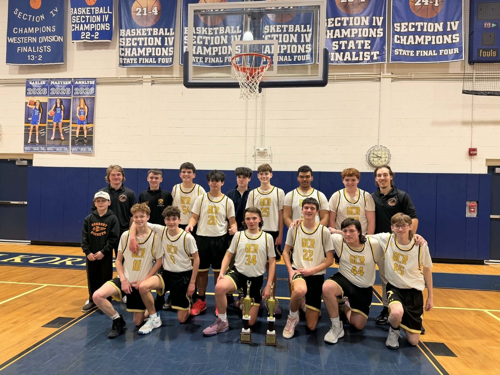 The image shows the GCR JV boys basketball team posing together on a gym floor. Some players are standing in the back row while others kneel in the front. They are wearing white jerseys with gold and black accents and “GCR” across the front. Two gold championship trophies are placed on the floor in front of the kneeling players.  Coaches stand at either end of the group. Behind them are blue championship banners hanging on the wall and a basketball hoop centered above the team. The players are smiling and leaning toward each other, capturing a proud moment after winning their championship.