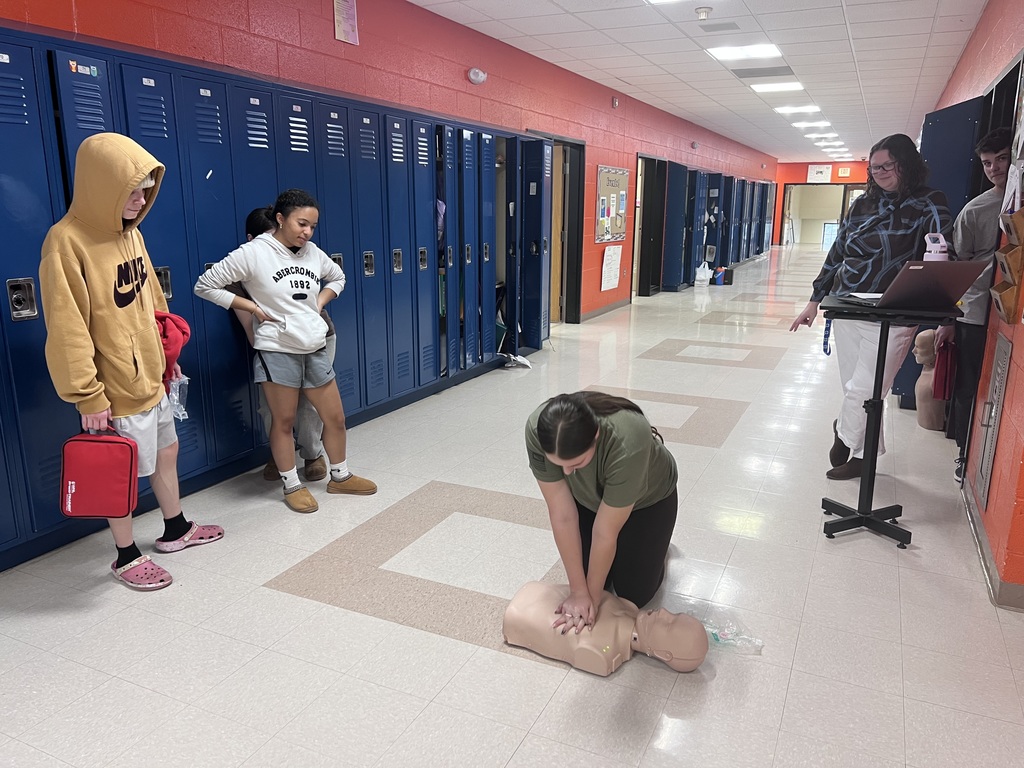 The image shows a group of high school students in a school hallway with blue lockers lining the wall and orange painted walls above them. The floor is a light colored tile, and the hallway stretches into the distance.  In the center of the image, a student kneels on the floor practicing CPR on a life sized training mannequin. The student’s hands are placed on the mannequin’s chest, demonstrating chest compressions.  Several other students stand nearby watching the demonstration. One student wearing a yellow hoodie and holding a red bag stands near the lockers. Another student in a light colored sweatshirt stands with hands on their hips observing the activity.  On the right side of the hallway, an adult instructor stands next to a small rolling cart with a laptop on top, appearing to supervise or guide the training. A mannequin head used for training equipment is also visible nearby.  The scene shows students actively participating in a CPR training exercise in a school hallway environment.