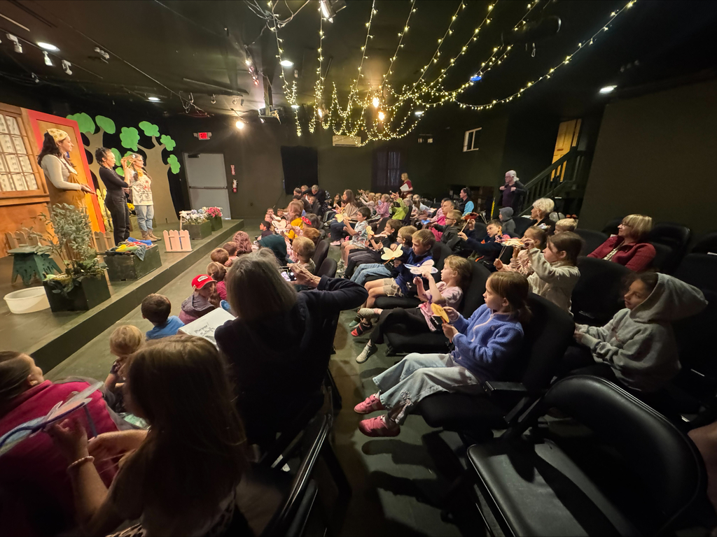 The photo shows a small theater filled with young elementary students seated in rows of dark chairs while watching a live stage performance. The room is dimly lit, with warm string lights hanging across the ceiling, creating a cozy and magical atmosphere. At the front of the theater, three actors stand on a colorful stage decorated with large flowers, trees, and a house backdrop. One performer appears to be speaking while the others interact nearby. Many students in the audience are holding paper butterfly crafts, raising them excitedly toward the stage as they participate in the show. The children, ranging from preschool through early elementary age, are dressed casually in sweaters, hoodies, and bright-colored clothing. Teachers and adults are seated among the students, helping supervise and enjoy the performance. The overall scene captures a joyful and engaging theater experience filled with curiosity, learning, and excitement.