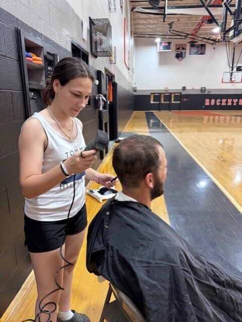 Gabby Miraglio stands behind Anthony Camillone who is seated in a chair with a barber shop cover over him. Gabby is holding barber sheers and is giving Anthony Camillone a hair cut.